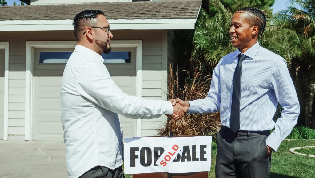 Two men shaking hands in front of house sold sign, sealing real estate deal outdoors. seo for real estate
