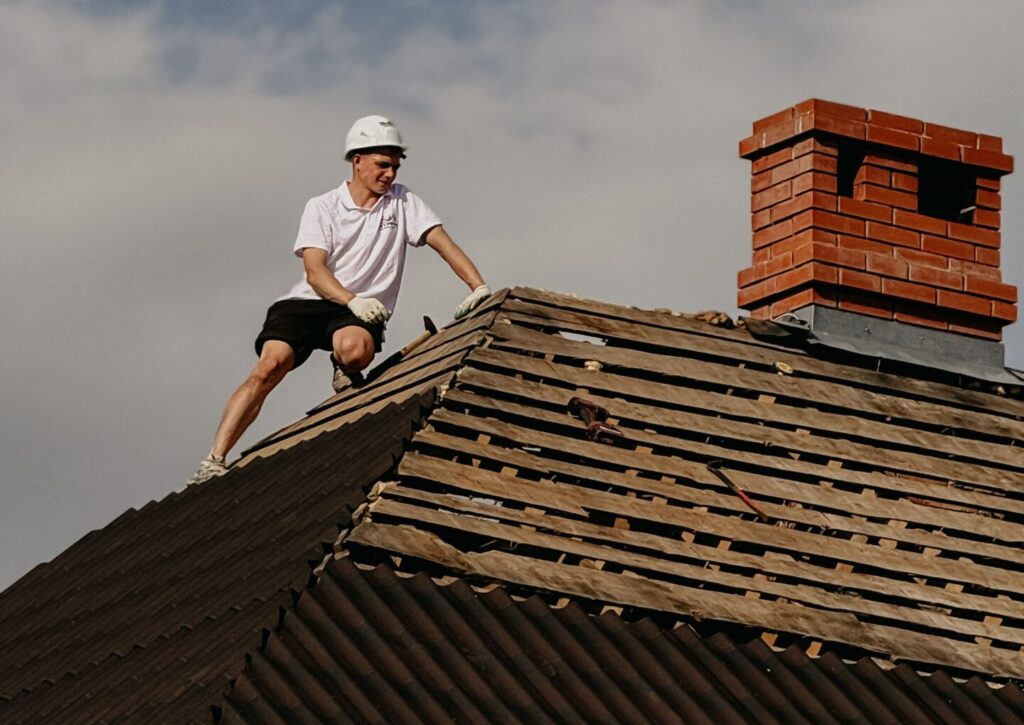pexels-photo-37623618-37623618 Man in safety helmet repairing roof of red brick house under clear sky.